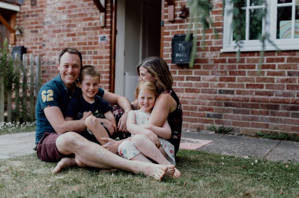 Family of four is sitting on the grass in front of their home. Family doorstep photo sessions in Hampshire. Ewa Jones Photography