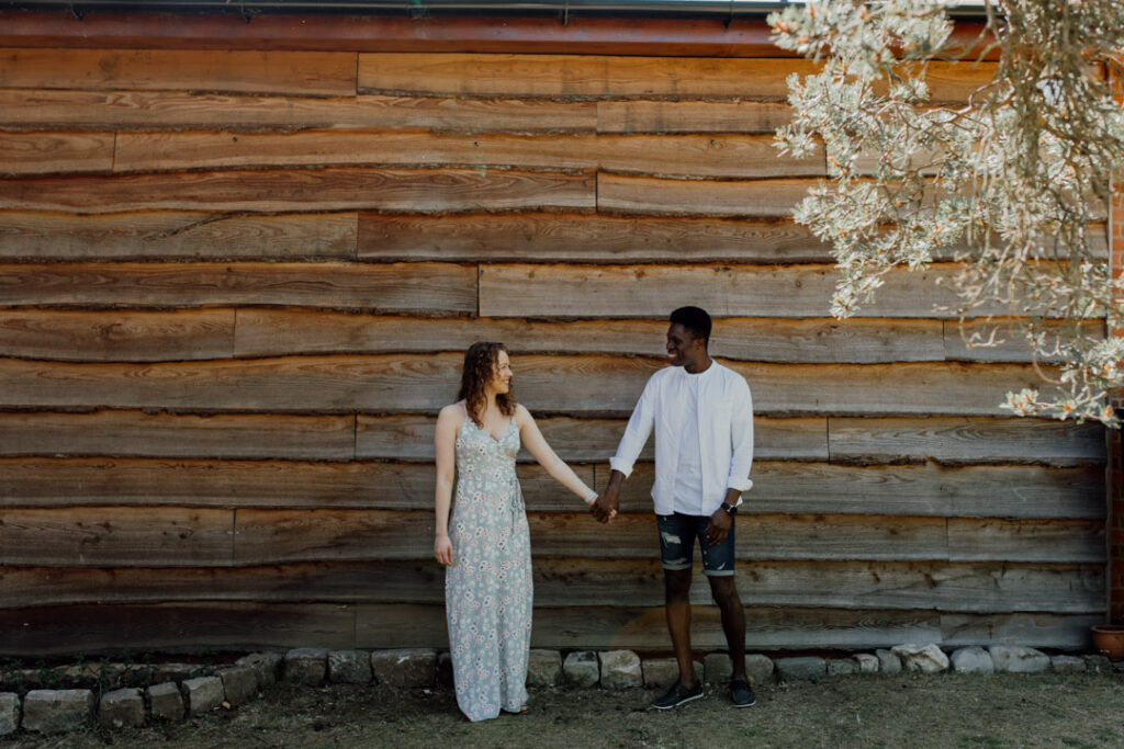 Engaged couple are standing and holding hands. They are standing in front of large wooden building. Looking at each other. Engagement photo session in Basingstoke. Ewa Jones Photography