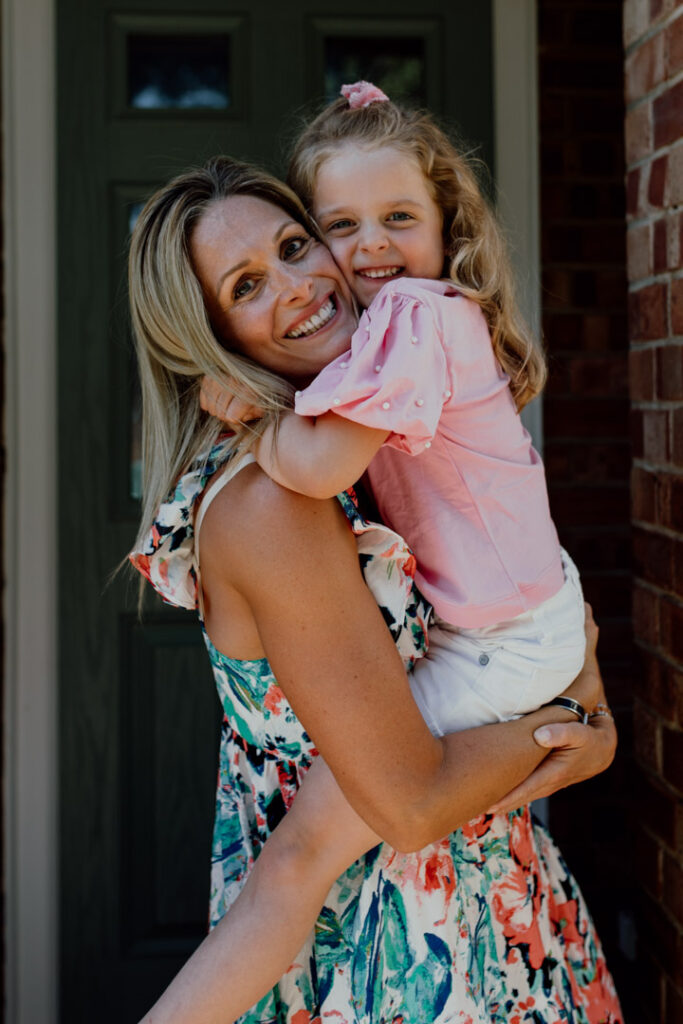 Mum is holding her daughter and both are smiling. Mum is wearing lovely colorful dress and girl is wearing pink top and white shorts. Family photographer in Basingstoke. Ewa Jones Photography