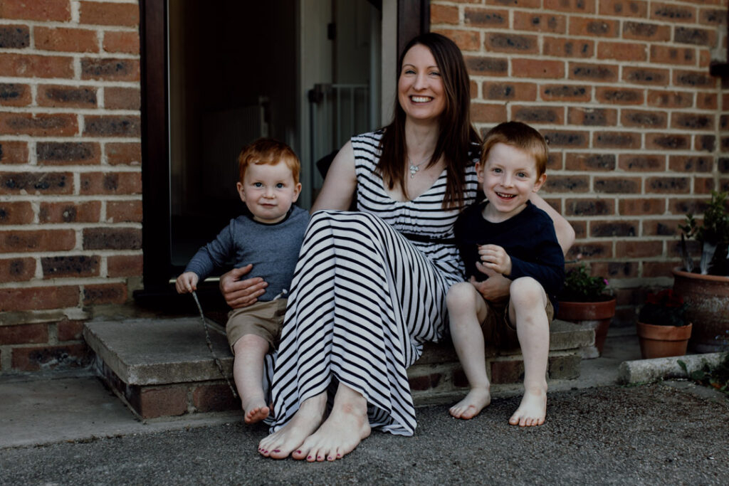 Mum is sitting on the doorstep and cuddling to her two boys. All are smiling. Family photography in Hamsphire. Ewa Jones Photography