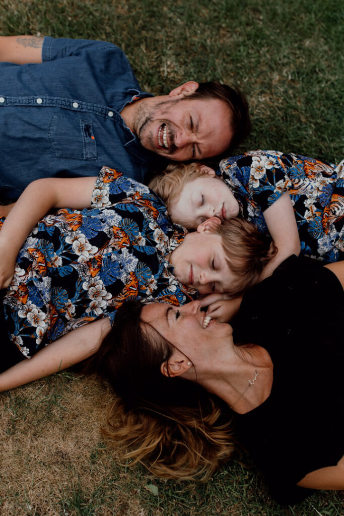 Mum, dad and two sons are laying on the grass. Mum and dad are looking at boys and both boys have their eyes closed. What to wear for a family photo session. Ewa Jones Photography