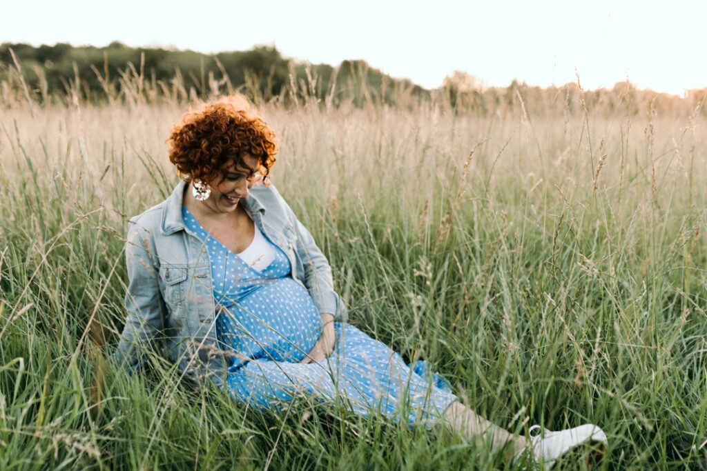 Pregnant mum is sitting on the long grass and laughing. She is wearing lovely blue dress with white spots and jean jacket. Maternity photography in Hampshire. Ewa Jones Photography