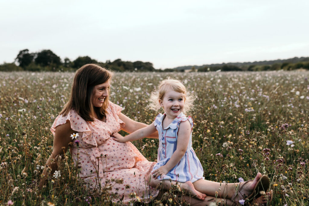 Mum and daughter are sitting on the grass. Baby kicked inside mummy tummy and little girl has a surprised face expression. Ewa Jones Photography