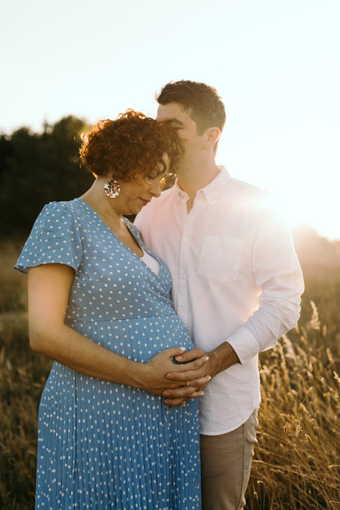 Expecting mum is holding her bump and husband is kissing mum on her head. Golden hour maternity photography in Hampshire. Ewa Jones Photography