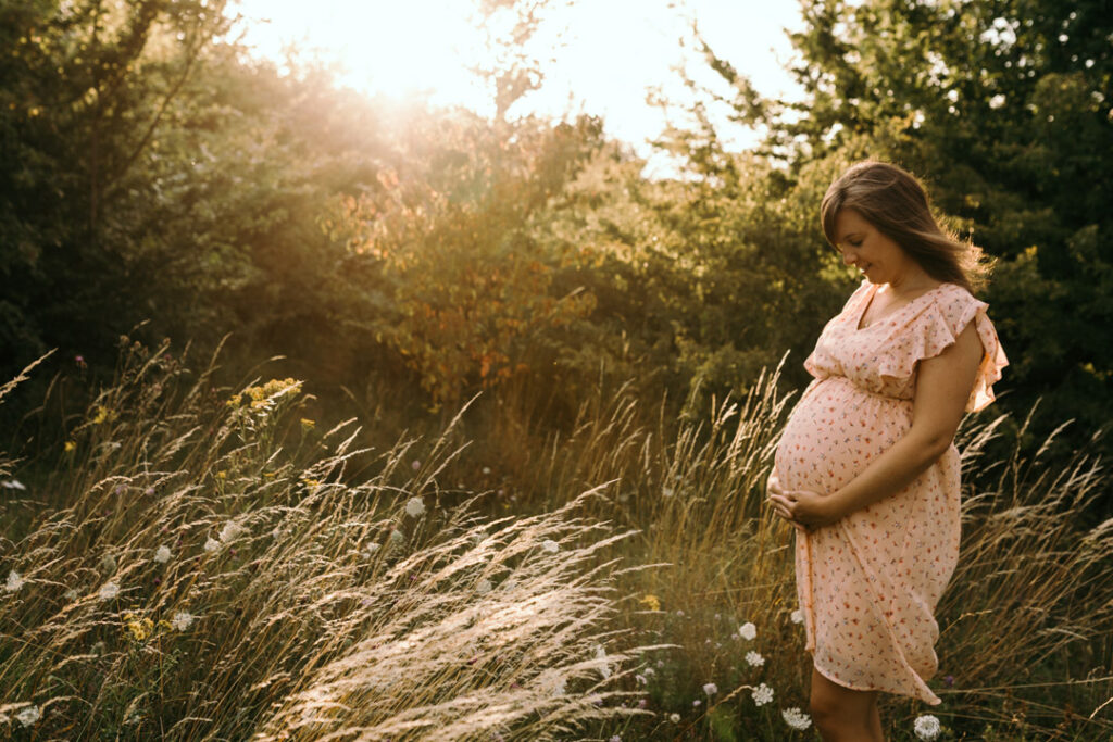 Expecting mum is holding her bump and looking down. She is standing in a lovely field. Sunset maternity photography in Hampshire. Ewa Jones Photography