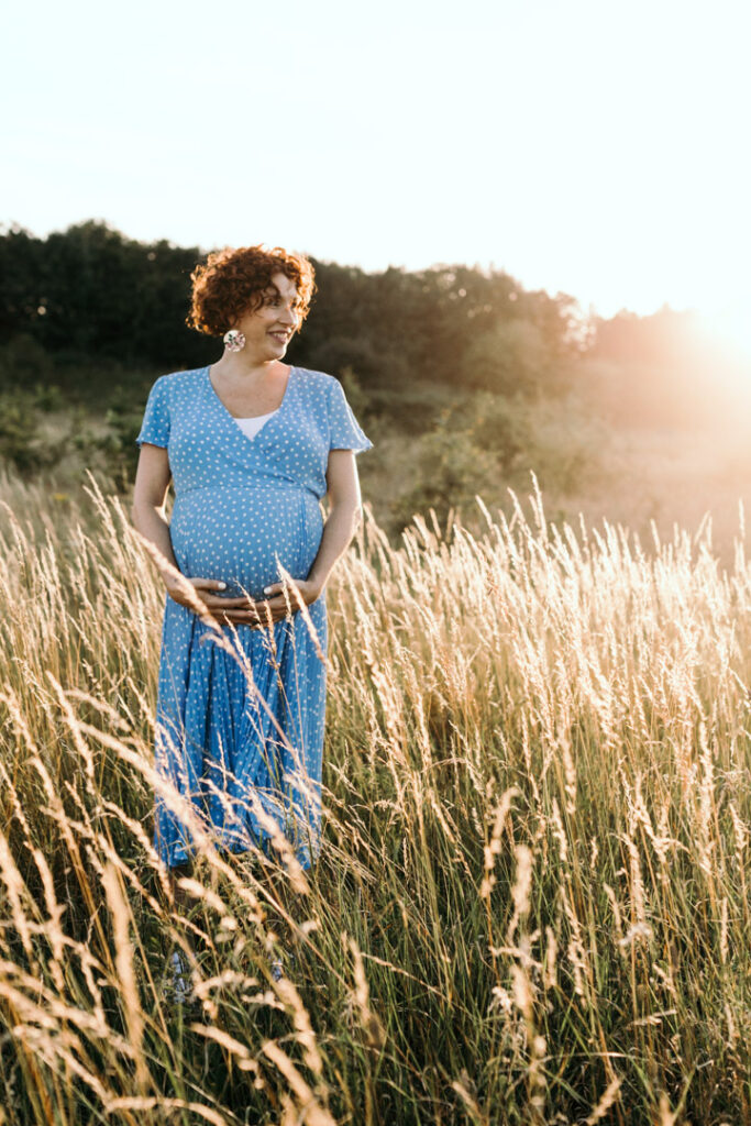 Expecting mum is wearing lovely blue dress with white dots. she is standing in the long grass. Maternity photography in Hampshire. Ewa Jones Photography