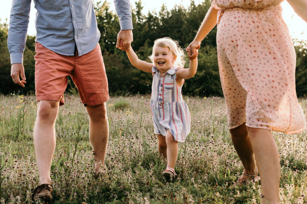 Expecting mum and dad are holding hands with their older daughter. The older daughter is laughing and running in the field of wild flowers. Maternity photo session in Hampshire. Ewa Jones photography