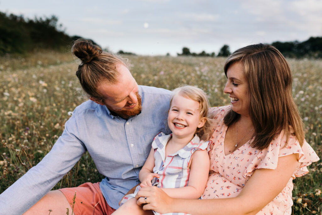 Pregnant mum is sitting with her partner and their older daughter. Mum and dad are looking at the daughter and laughing. Mum is wearing lovely pink dress, daughter is wearing stripy dress and dad is wearing blue shirt and red trousers. Lovely sunset maternity photo session in Basingstoke. Ewa Jones Photography