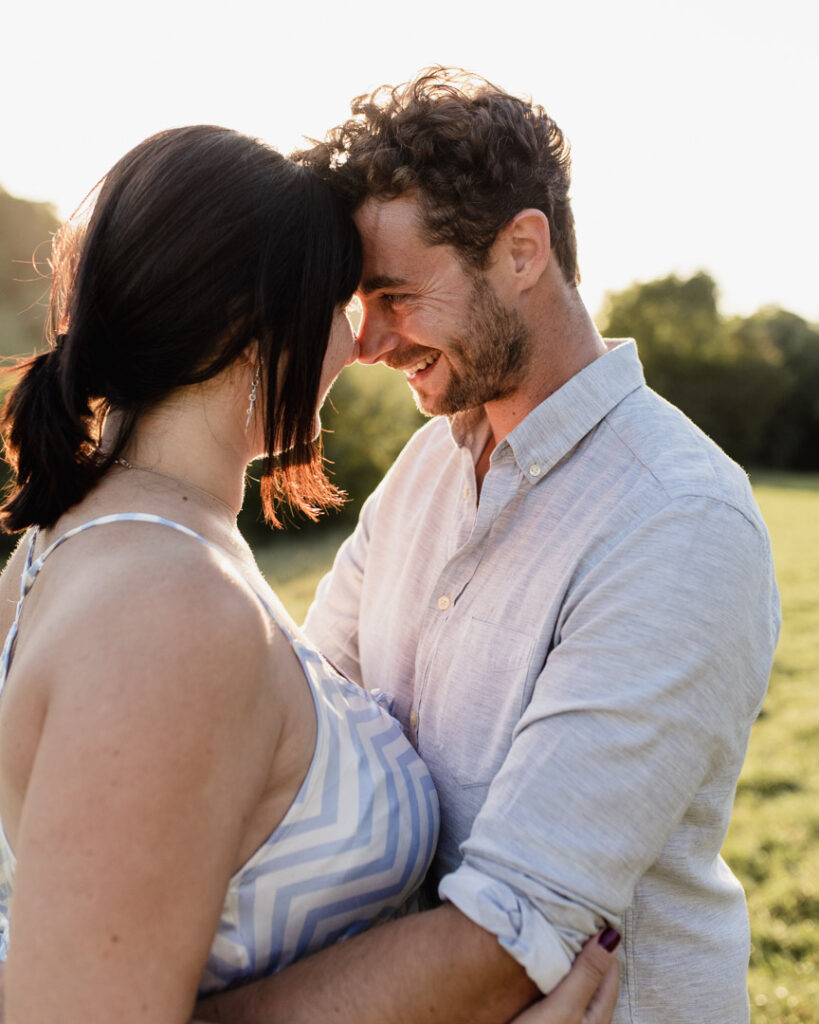 Dad is looking lovingly at his partner. Both are touching foreheads and smiling to each other. Sunset maternity photo session. Ewa Jones Photography