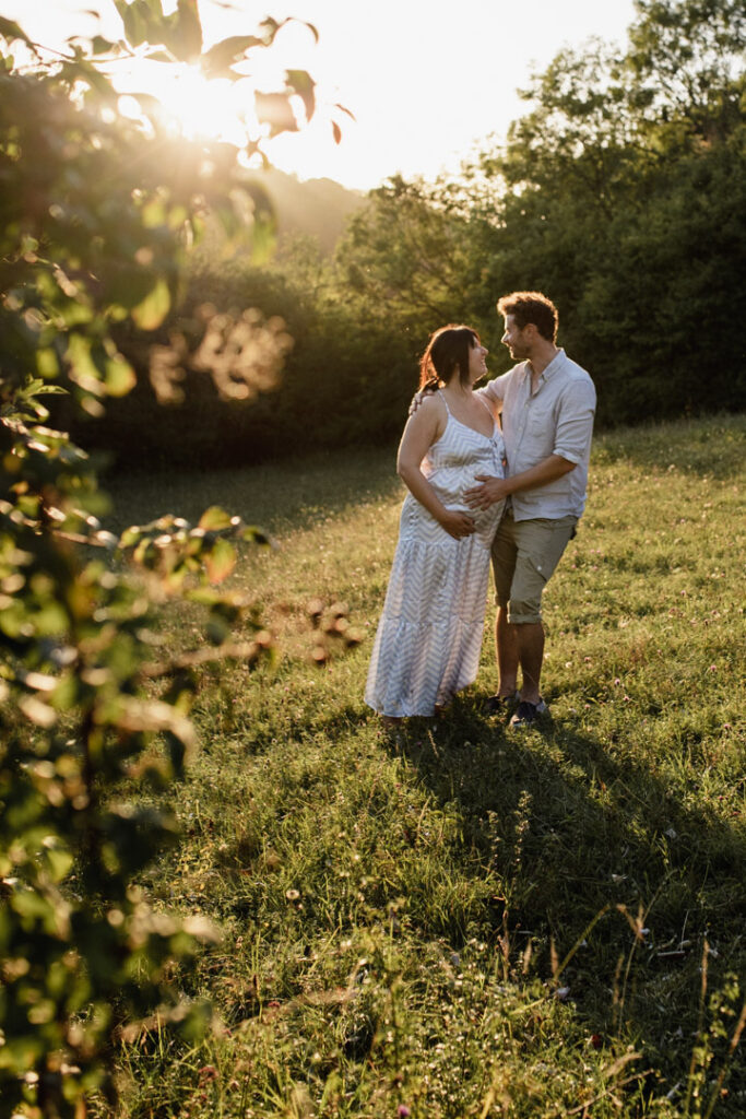 Expecting parents are standing in the fields and looking at each other. The sun is shining. Lovely maternity photo session. Pregnant mum is sitting with her partner and their older daughter. Mum and dad are looking at the daughter and laughing. Mum is wearing lovely pink dress, daughter is wearing stripy dress and dad is wearing blue shirt and red trousers. Lovely sunset maternity photo session in Basingstoke. Ewa Jones Photography
