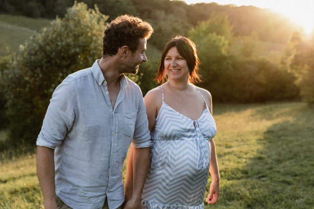 Pregnant mum is walking with her partner and holding hands. She is wearing lovely dress with patterns and her partner is wearing grey shirt. What to wear for a maternity photo session. Ewa Jones Photography