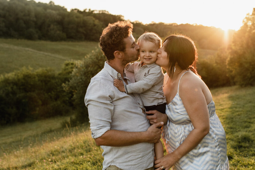 Expecting mum is kissing her toddler boy and dad is kissing his boy. Mum is wearing lovely maternity dress and dad and boy are wearing light blue shirts. What to wear for a maternity photo session. Ewa Jones Photography