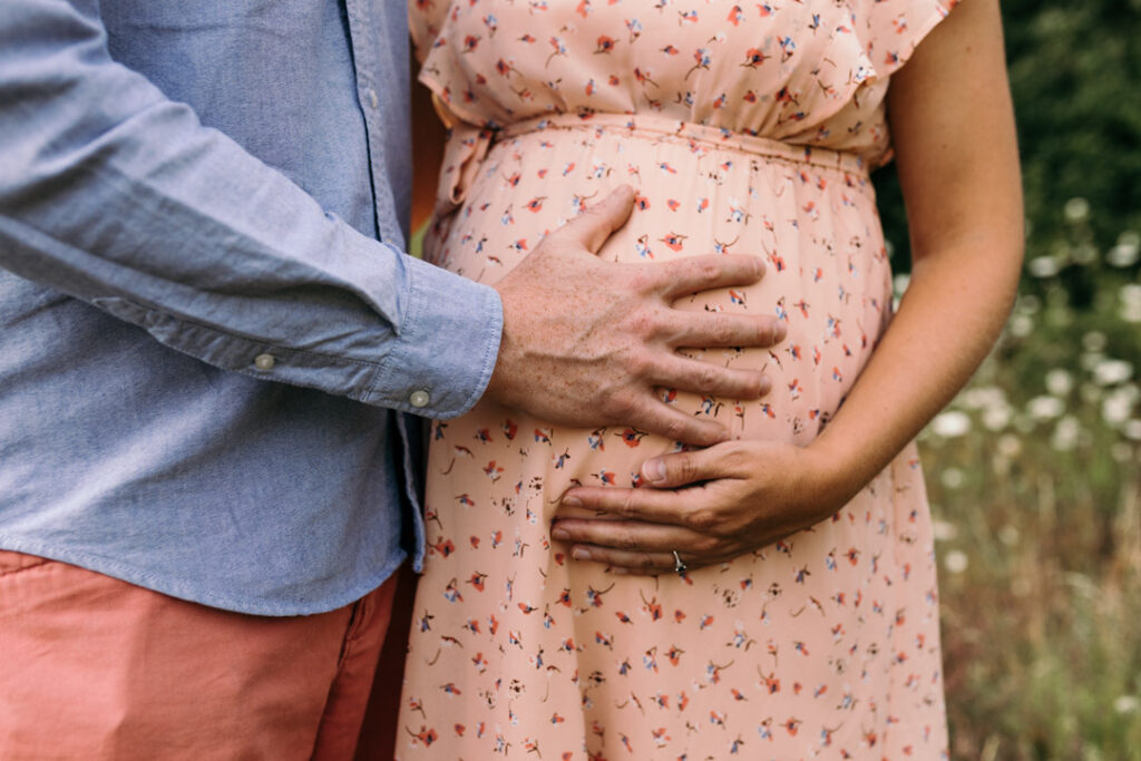 Dad and mum are holding hands on the bump. Mum is wearing lovely salmon colour dress with flowers. Maternity photography in Hampshire. Ewa Jones Photogarphy
