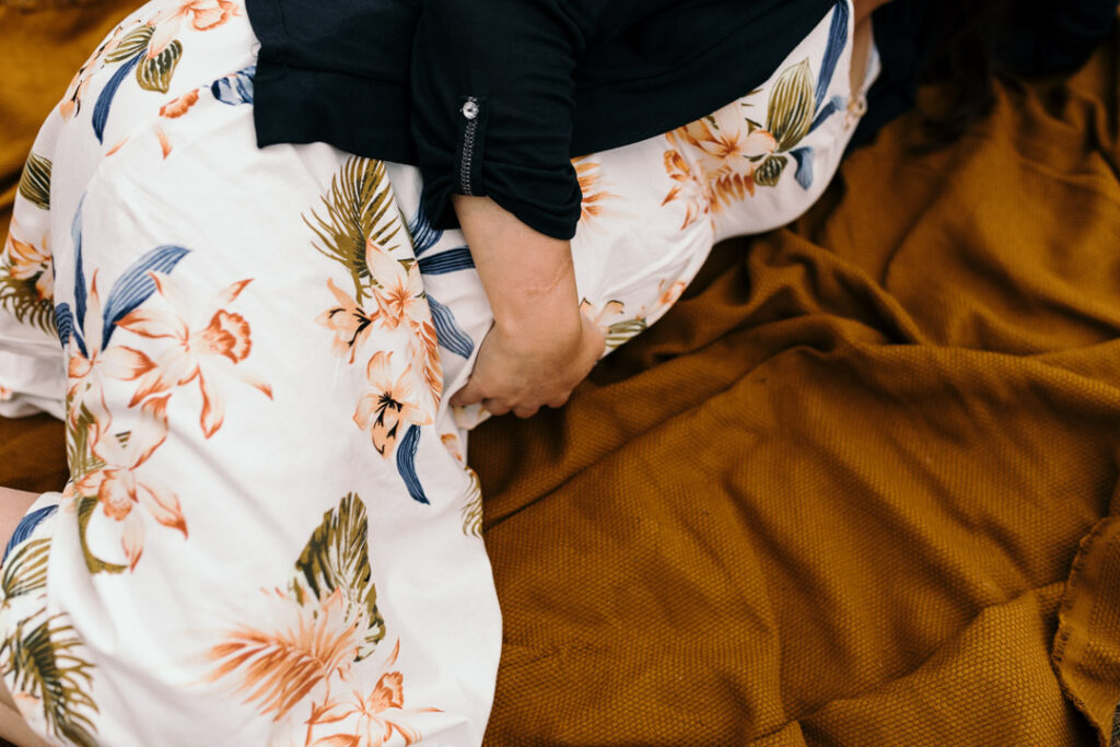 Pregnant mum is laying on the dark orange blanket and holding her bump. She is wearing lovely white dress with flowers. What to wear for a maternity photo session. Maternity photography in Hampshire. Ewa Jones Photography