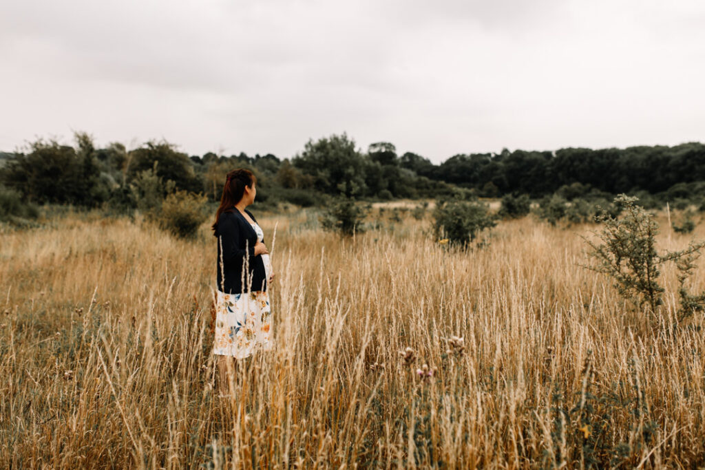 Expecting mum is standing in the long grass and looking away. She is wearing cream dress with flowers and dark blue cardigan. Maternity photo session in Hampshire. Ewa Jones Photography