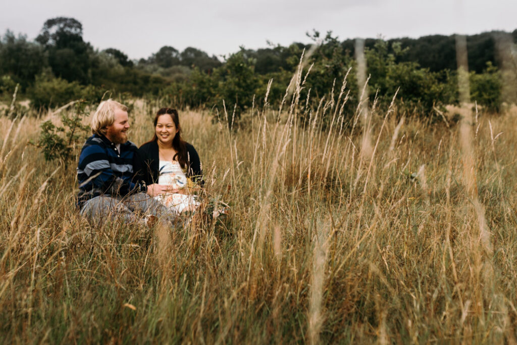 Expeciting parents are sitting on the grass and smiling. Pregnant mum is sitting with her partner and their older daughter. Mum and dad are looking at the daughter and laughing. Mum is wearing lovely pink dress, daughter is wearing stripy dress and dad is wearing blue shirt and red trousers. Lovely sunset maternity photo session in Basingstoke. Ewa Jones Photography