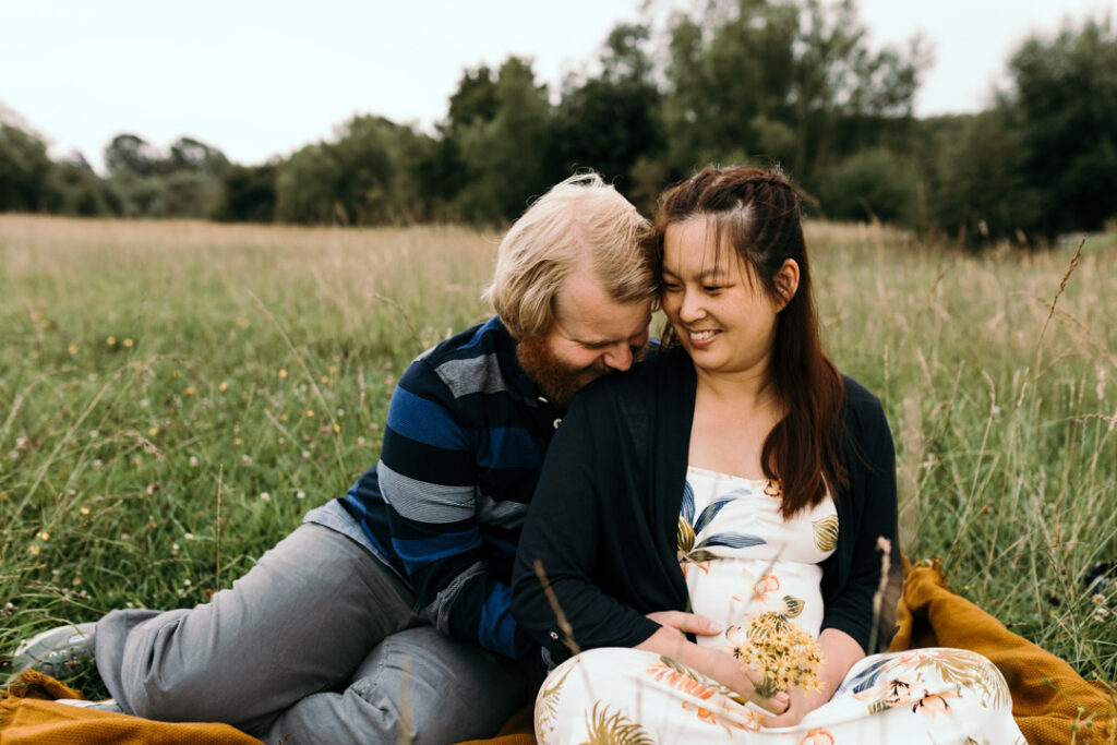 Expecting mum is sitting on the blanket with her husband. Her husband is laughing and holding her bump. Mum is wearing lovely cream dress with flower patterns. What to wear for a maternity photo session. maternity photography in Basingstoke, Hampshire. Ewa Jones Photography