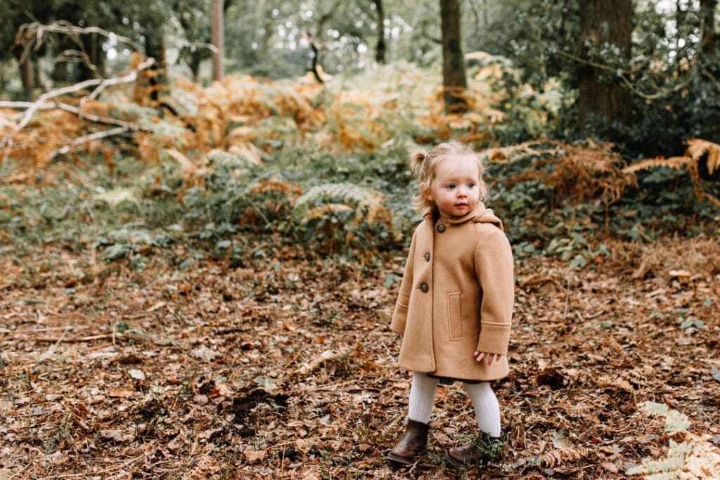 Girl is standing in the woods and wearing lovely brown coat, white tights and brown shoes. Autumn family photo sessions in Basingstoke. Ewa Jones Photography
