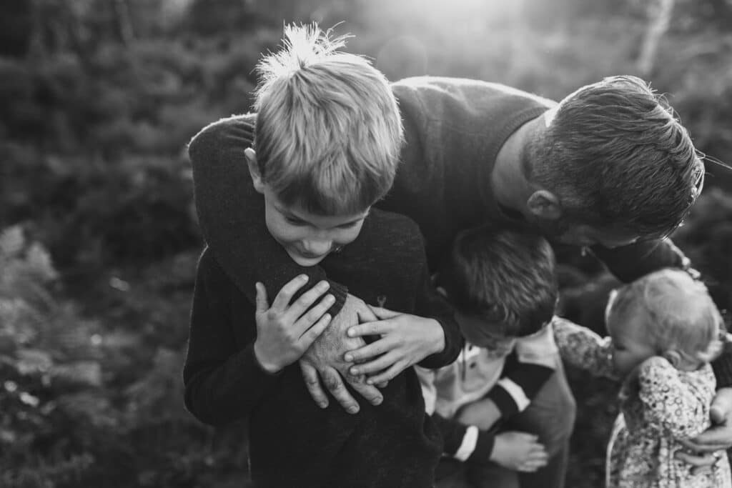 Dad is holding his three children and one boy is holding onto dads hand. Lovely black and white lifestyle family photography in Hampshire. Ewa Jones Photography