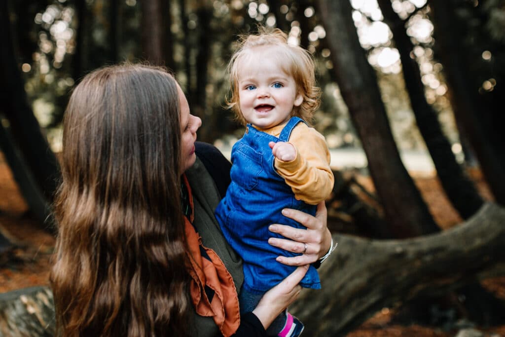 Mum is holding a little girl and looking at her. Girl is smiling and she is wearing a blue jean dress and yellow top. Family autumn session. Ewa Jones Photography