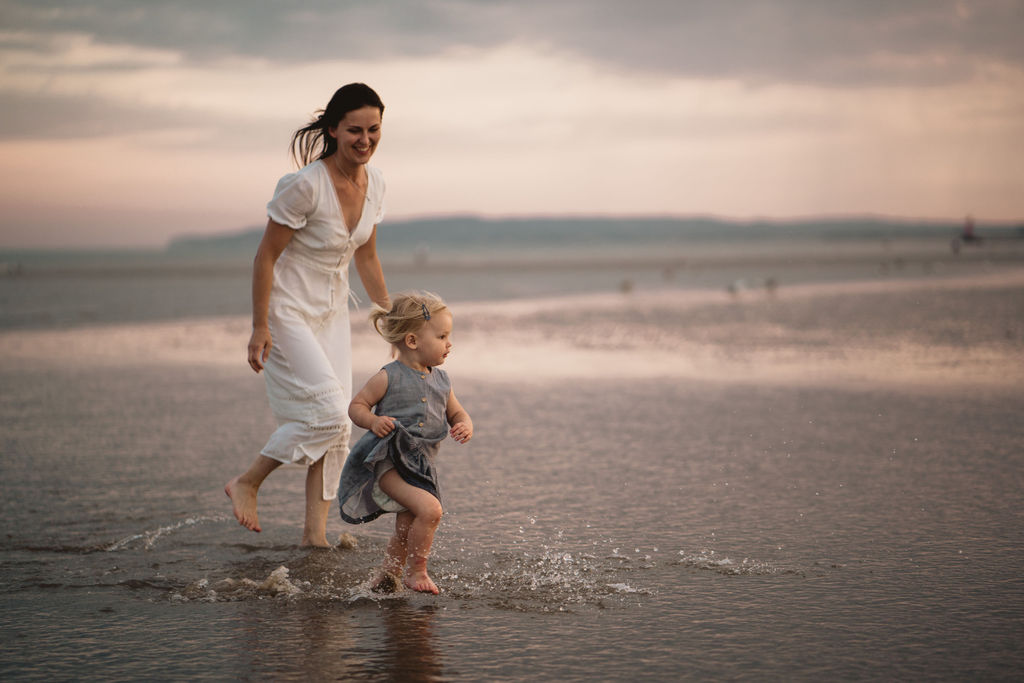 Mum is chasing little girl. Mum is wearing a white dress and little girl is wearing a grey dress. Our first family photo session
