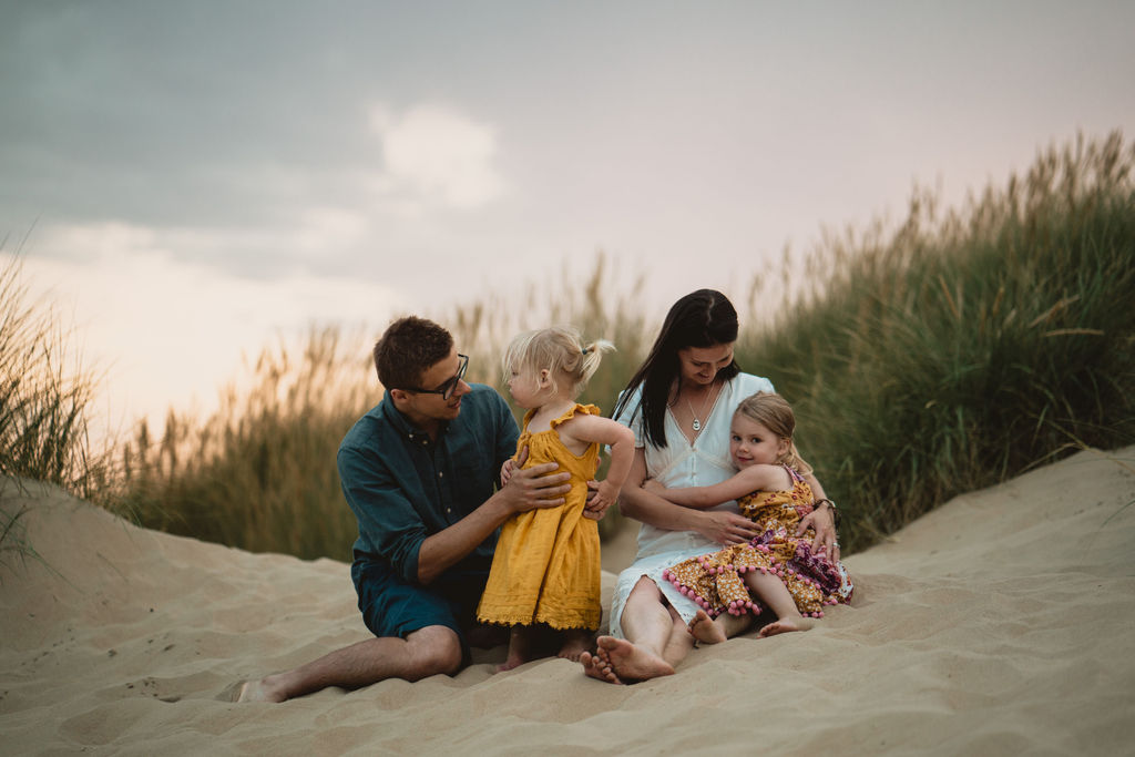 Family of four is sitting on the sand dunes and cuddling to each other. Lovely candid family sunset photography