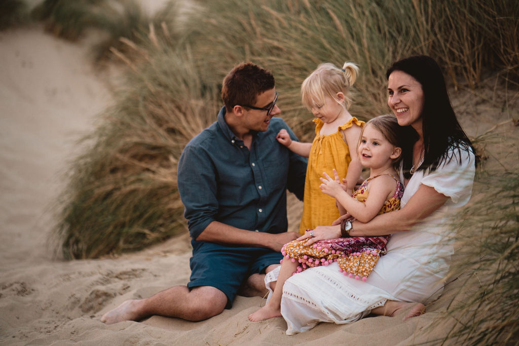 Family of four is sitting on the sand dunes and cuddling to each other. Lovely candid family sunset photography