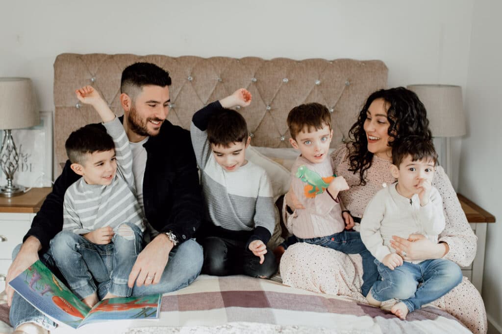 Family of six is sitting on the bed and looking at each other. Two boys are raising their hands and mum and dad are lovingly watching them. Family photography in Hampshire. Ewa Jones Photography