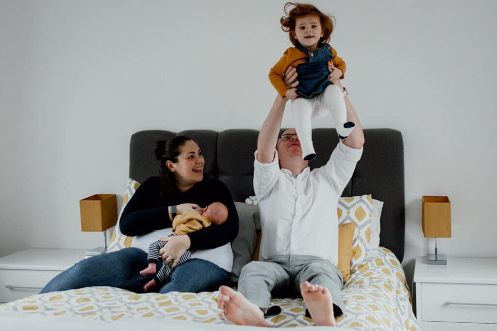 Mum, dad, older sister and little newborn are on the bed. Mum is looking up at the older daughter laughing when dad is throwing girl into the air. Lifestyle newborn photography in Hampshire. Ewa Jones Photography