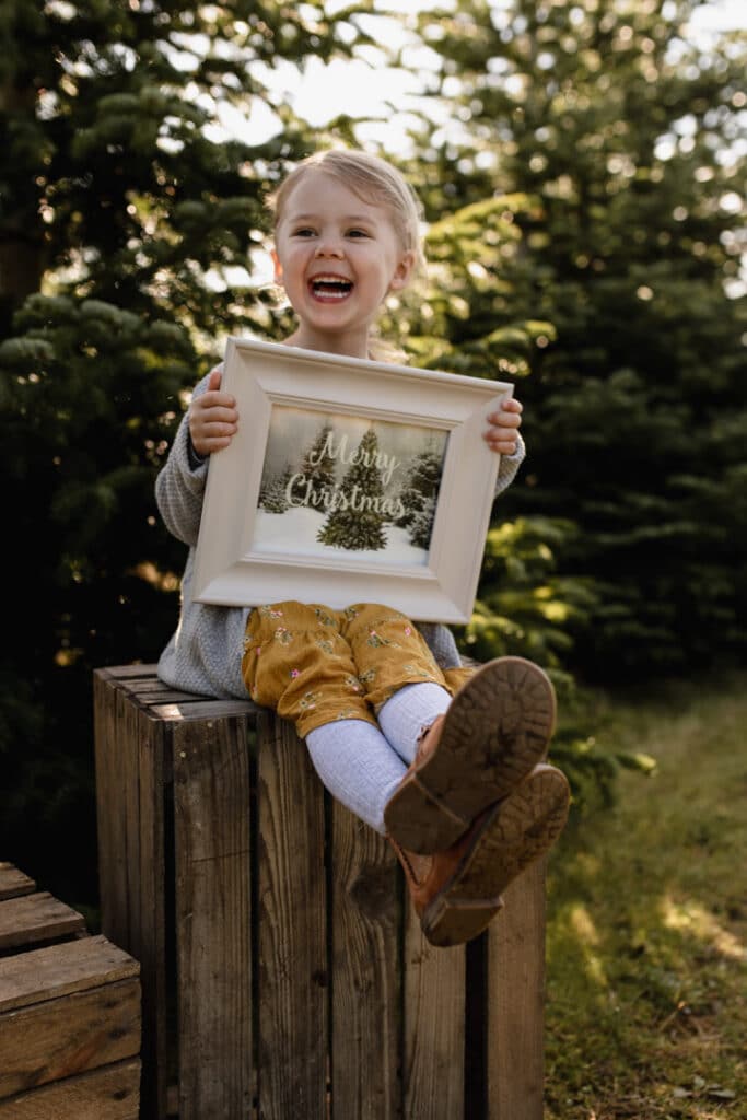 Little girl is sitting on the apple crate and holds frame with merry christmas wording. She is smiling and is on the Christmas tree farm. Family Christmas sessions. Ewa Jones Photography 