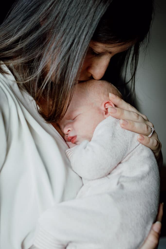 Mum is holding her newborn baby very close to her chest and kissing her. Newborn baby is sleeping. Newborn baby photographer in Basingstoke, Hampshire. Ewa Jones Photography