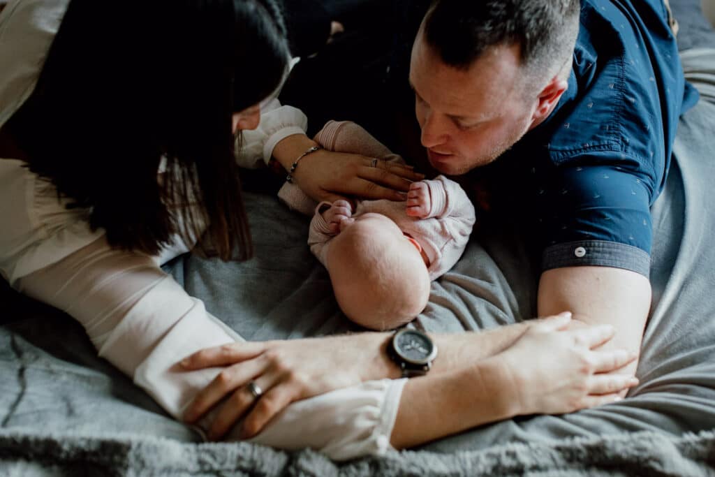 Mum and dad are laying on bed and interlocking their hands together. They are looking at their newborn baby girl. Newborn photographer in Basingstoke, Hampshire. Ewa Jones Photography