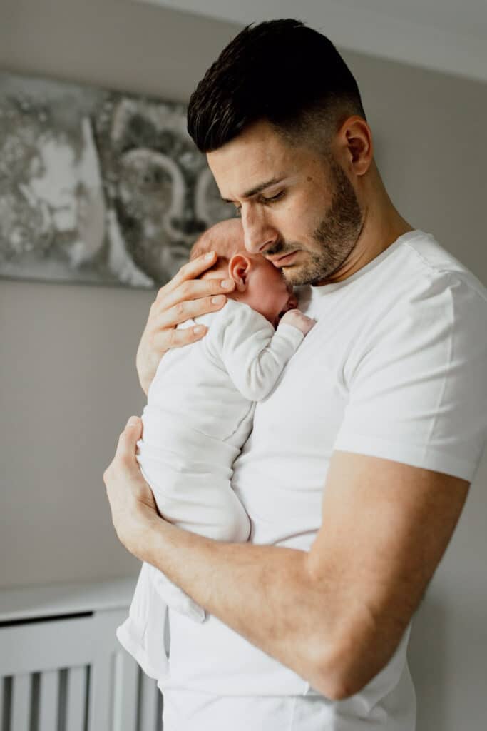 Dad is holding his newborn baby close to his chest. He is standing close to the window. Newborn baby photography in Basingstoke, Hampshire. Ewa Jones Photography