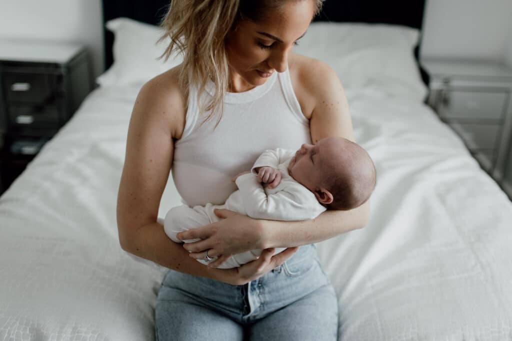 Mum is holding her newborn baby who is sleeping. She is sitting on the edge of the bed. Mum is wearing white top and blue jeans. Baby is wearing white baby grow. What to wear for your newborn baby photo shoot. Newborn baby photography in Basingstoke, Hampshire. Ewa Jones Photography