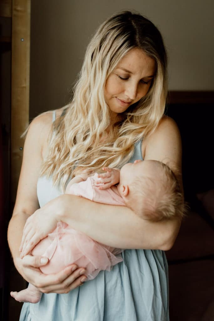Mum is holding her newborn baby girl close to her and looking at her. Mum is wearing blue dress and her newborn baby girl is wearing pink baby grow. What to wear for your newborn baby photo shoot. Newborn baby photographer in Hampshire. Ewa Jones Photography