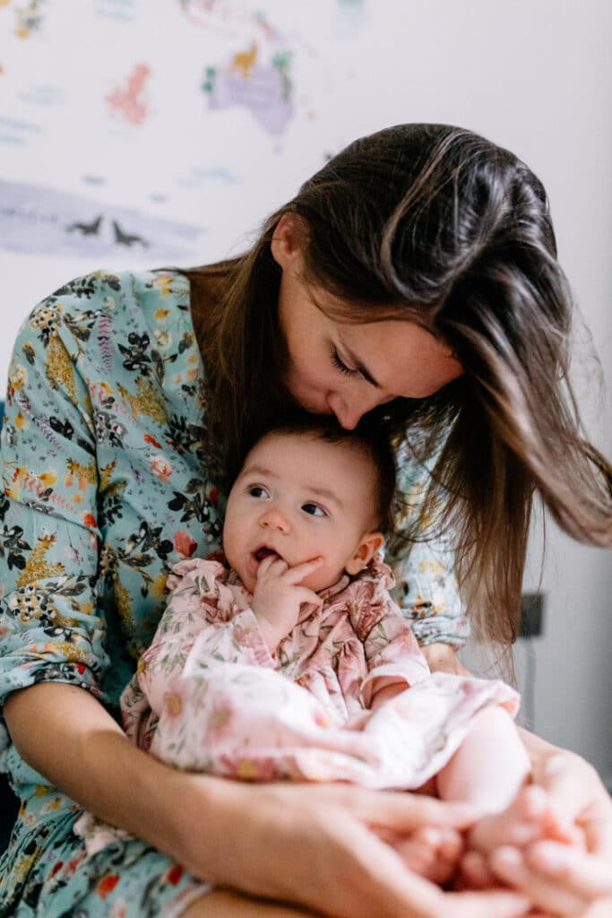 Mum is sitting on the bed and kissing his little baby girl. She is wearing a lovely green dress with lots of flowers and her little baby girl is wearing pink dress. What to wear for your newborn baby photo shoot. Newborn photography in Hammersmith. Ewa Jones Photography