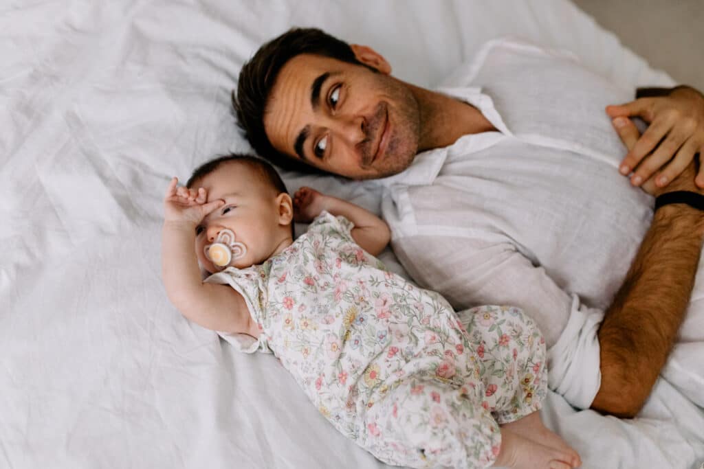 Dad is laying on bed and looking at his baby daughter. His daughter is laying next to her dad and wearing lovely flowery baby grow. What to wear for your newborn baby photo shoot. Newborn baby photographer in Hampshire. Ewa Jones Photography