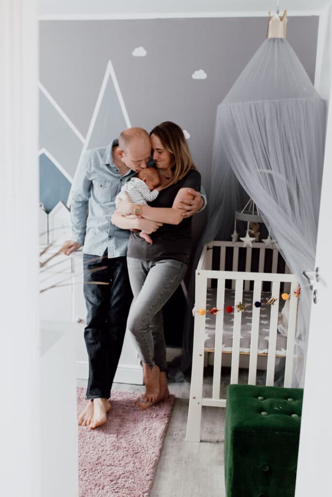 Mum and dad are standing in little baby girl room. Mum is holding newborn baby girl and dad is kissing his baby. They are all looking lovely and happy. Newborn photography in Basingstoke, Hampshire. Ewa Jones Photography