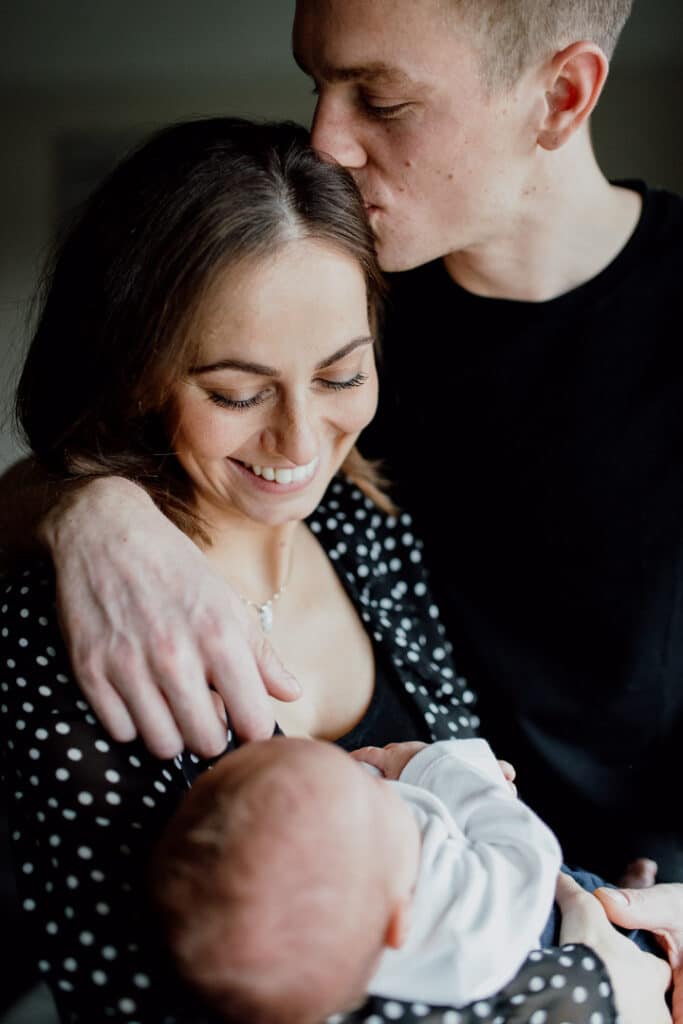 Mum and dad are standing close to each other and mum is holding a newborn baby. Dad is kissing mum on her head and mum is smiling. Newborn baby photographer in Hampshire. Ewa Jones Photography