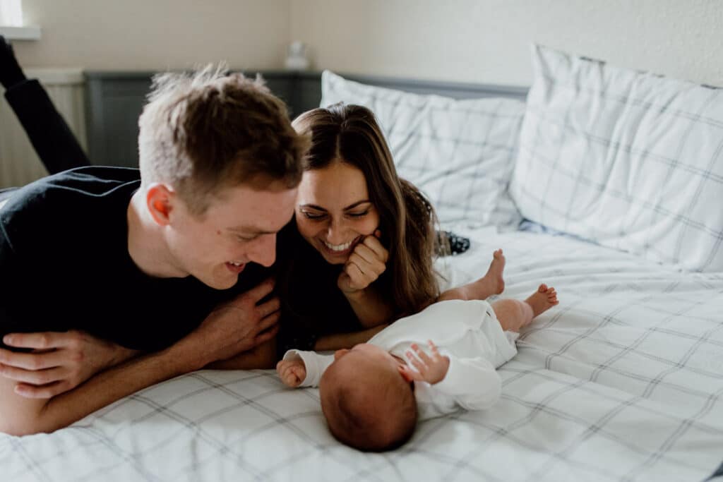 Mum and dad are laying on the bed and looking at their newborn baby. Mum is wearing black dress with white dots and dad is wearing black t-shirt. Newborn baby is wearing a white long sleeve baby grow. What to wear for your newborn baby photo shoot. Basingstoke, Hampshire. Ewa Jones Photography