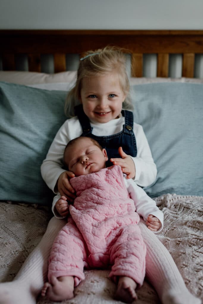 Older sister is sitting on the bed and holds her little newborn baby sister. Newborn baby is sleeping and wearing pink dungarees and older sister is wearing blue dress. Newborn photography in Basingstoke, Hampshire. Ewa Jones Photography