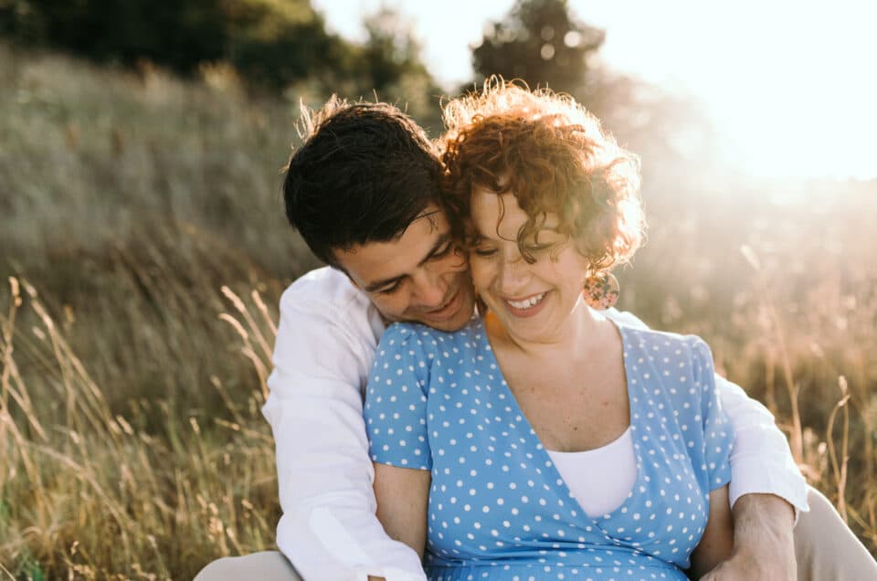 Pregnant mum is sitting on the grass and her husband is sitting behind her. they both look down and smiling. Mum is wearing lovely blue dress and dad is wearing white shirt. Why I love taking maternity photo sessions. Maternity photographer in Hampshire. Ewa Jones Photography