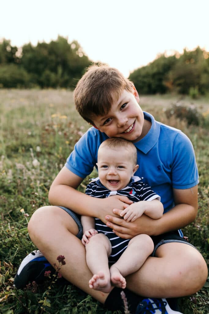 Older cousin is holding his little baby cousin on his lap. They are both laughing. Family photo session during golden hour. Family photographer in Basingstoke, Hampshire. Ewa Jones Photography