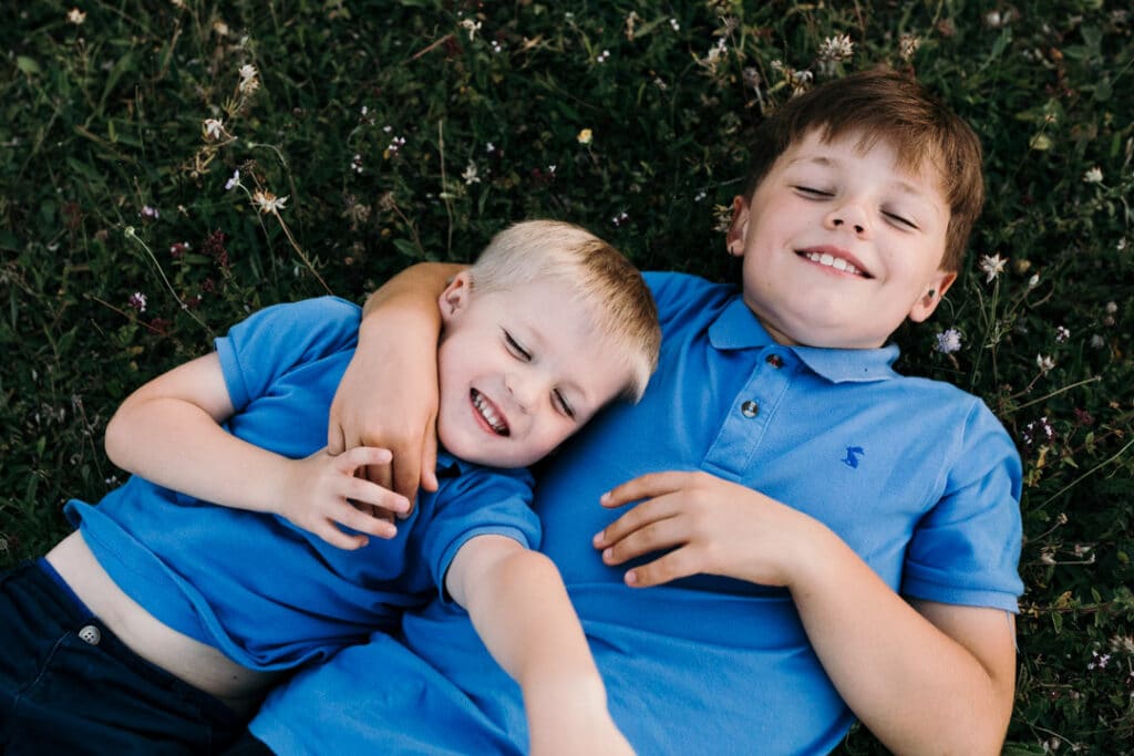 two cousins are laying on the grass and have eyes closed. They are both smiling. Both boys are wearing blue tops and shorts. family photographer in Hampshire. Ewa Jones Photography