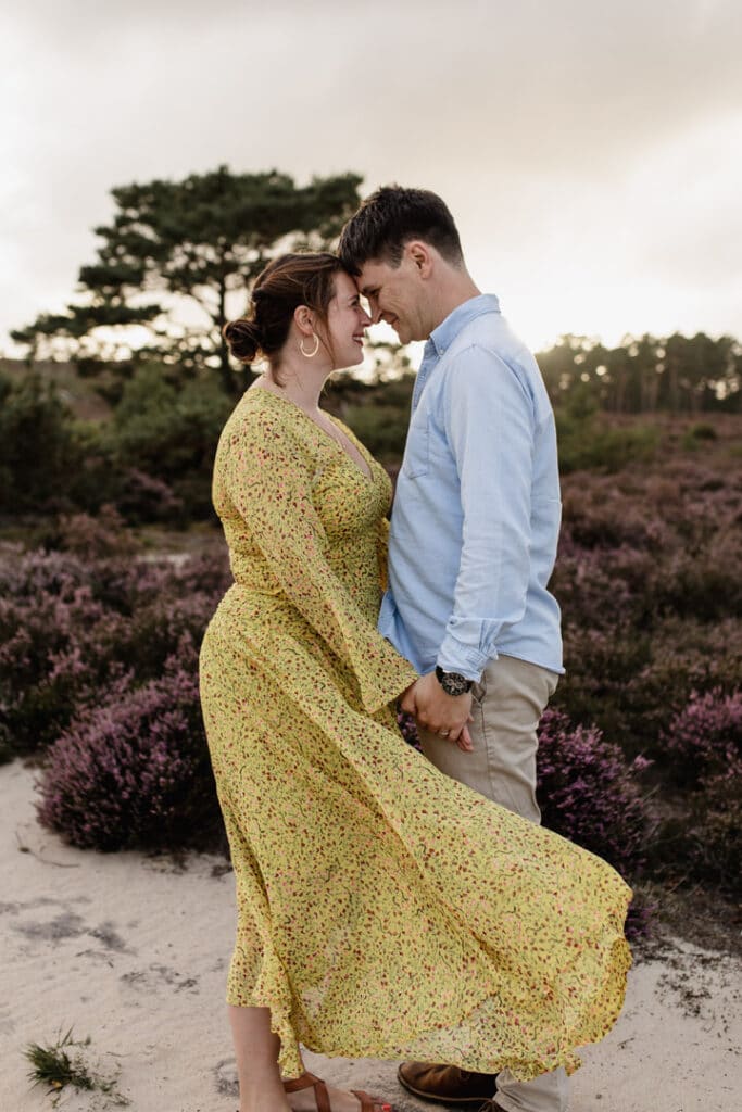 Mum and dad are holding hands together and are touching foreheads. Mum is wearing lovely yellow dress and dad is wearing blue shirt and light cream chinos. They are standing in the lovely common. Purple heather is on the ground. Family photographer in Hampshire. Ewa Jones Photography