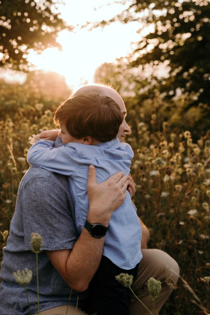 Dad is cuddling tight his son. Golden sun is shining behind dad and son. Documenting real family moments. Family photography in Hampshire. Ewa Jones Photography