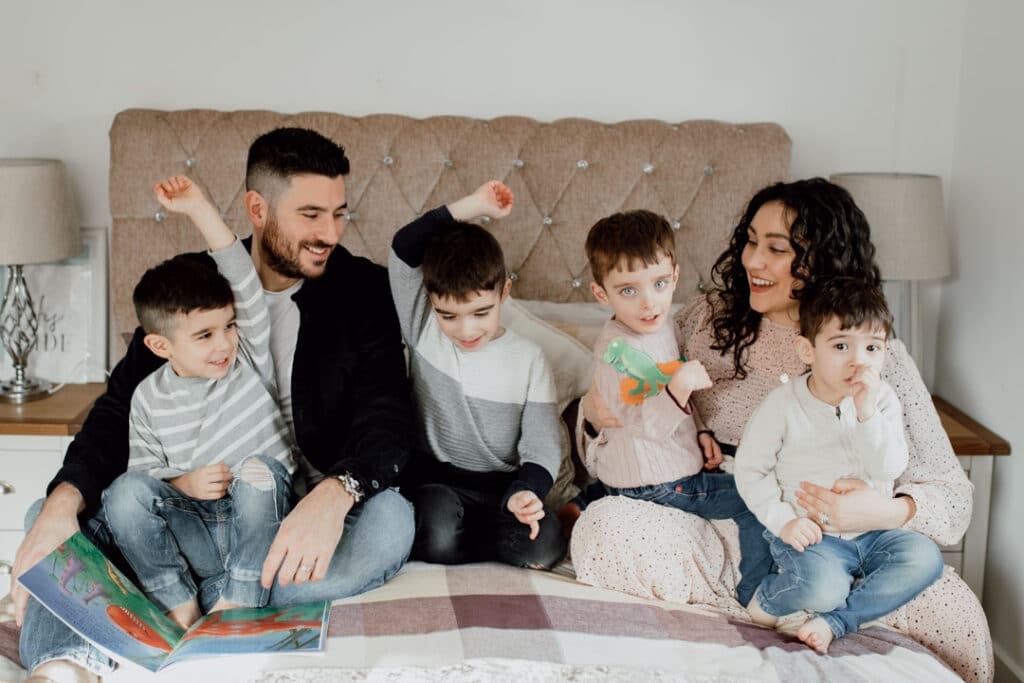 Family of six are all sitting on the bed and reading books. Mum is wearing lovely dress and her boys are wearing jeans and jumpers. All are smiling. Family photographer in Hampshire. Ewa Jones Photography