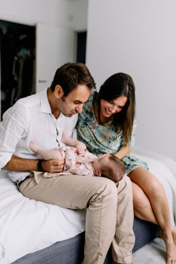 Mum and dad are sitting on the bed and looking at their baby daughter. Dad is making silly faces to her daughter. Mum is looking at the daughter and smiling. Family lifestyle photography. Ewa Jones Photography