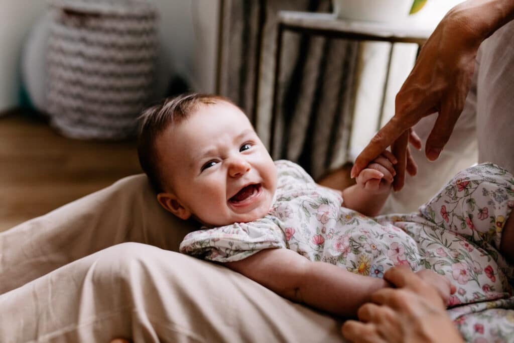 Little baby girl is looking at the camera and smiling. She is wearing lovely flowery outfit and dad is holding her hand. She is laying on dad's knees. family photographer in Hampshire. Ewa Jones Photography