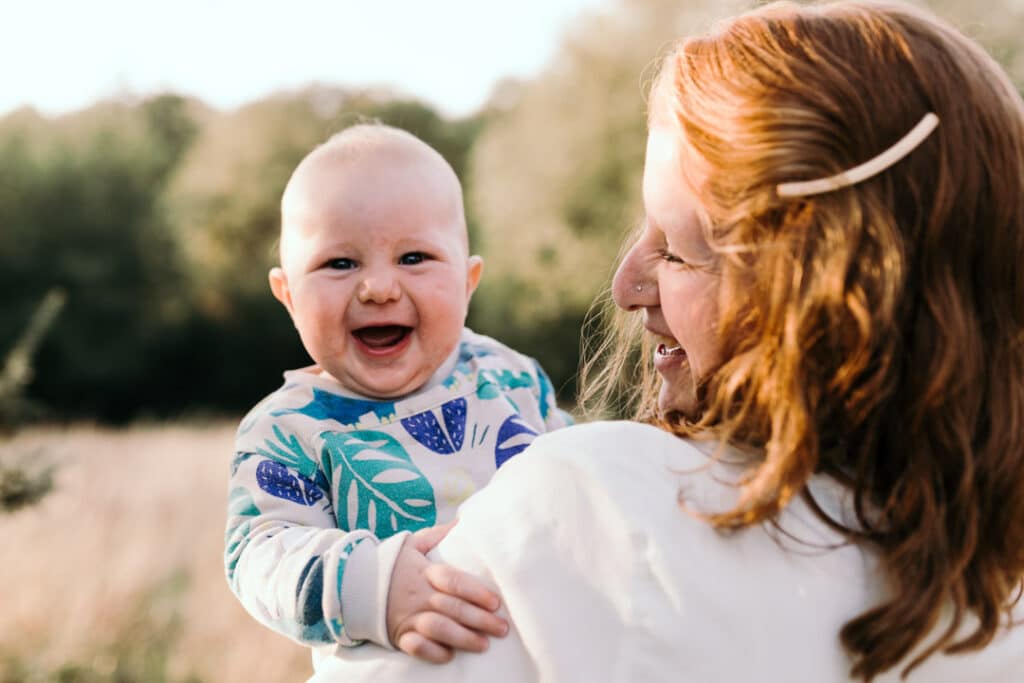 Mum is holding her six month old boy and looking at him. Sun is shining on mum's hair. Little boy is looking at the camera and smiling. Family photographer in Hampshire. Ewa Jones Photography